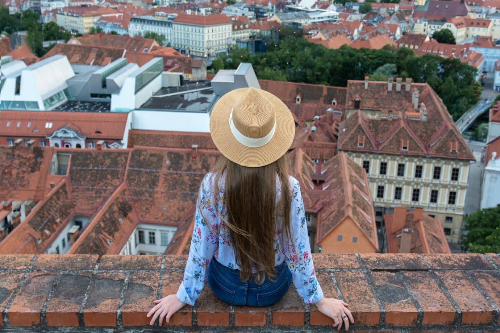 A girl with long hair in a hat sits on the edge of a tall building and looks at the city of Graz.