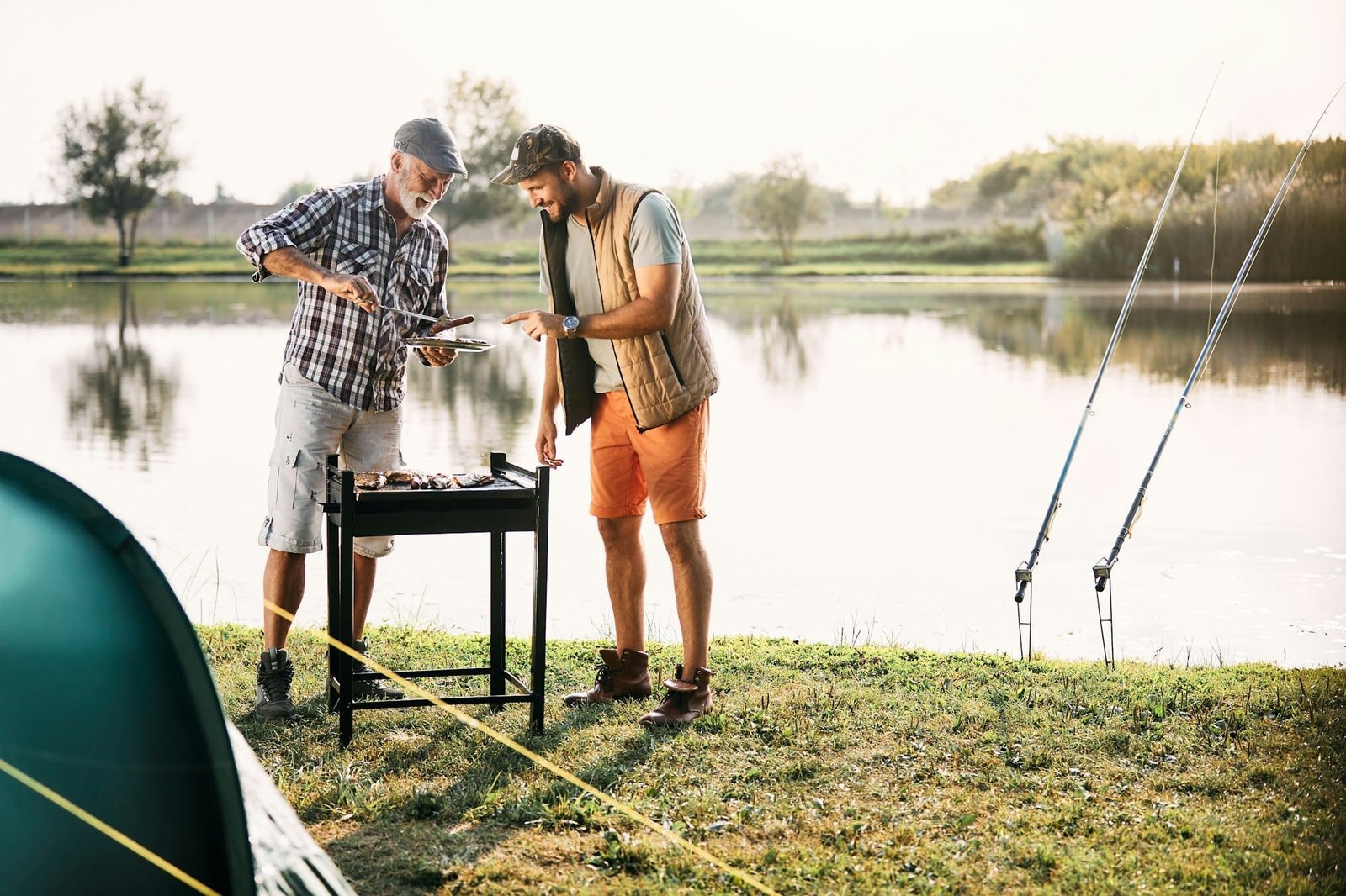 Happy mature man and his adult son grilling meat while camping by the lake.